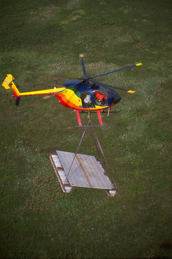 Helicóptero transportando plataforma metálica con eslinga sobre pastizal